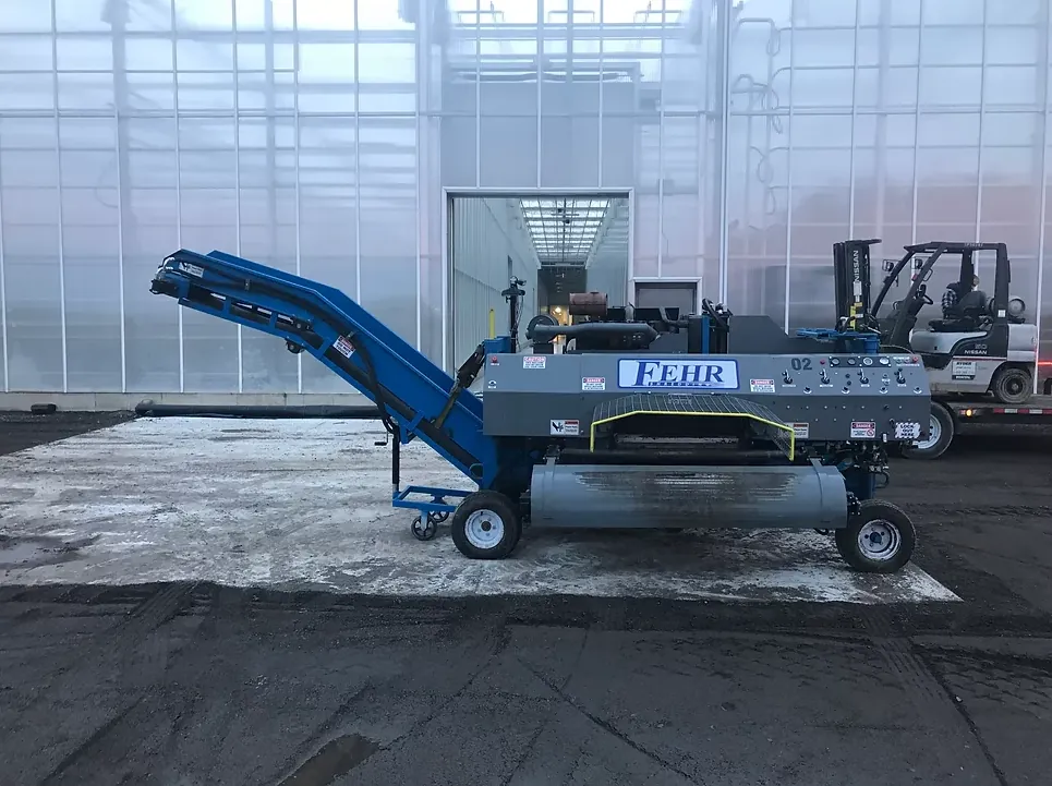 A blue Fehr shredding machine with its conveyor raised, parked outside a greenhouse